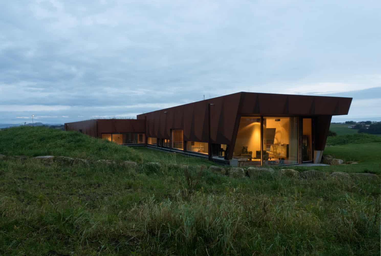 Contemporary modern house with angular rust-colored exterior on green landscape during dusk.