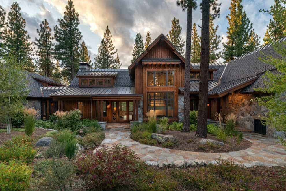 Modern mountain cabin with rustic wood exterior surrounded by tall pine trees and landscaped garden, showcasing contemporary architecture blended with natural forest elements.