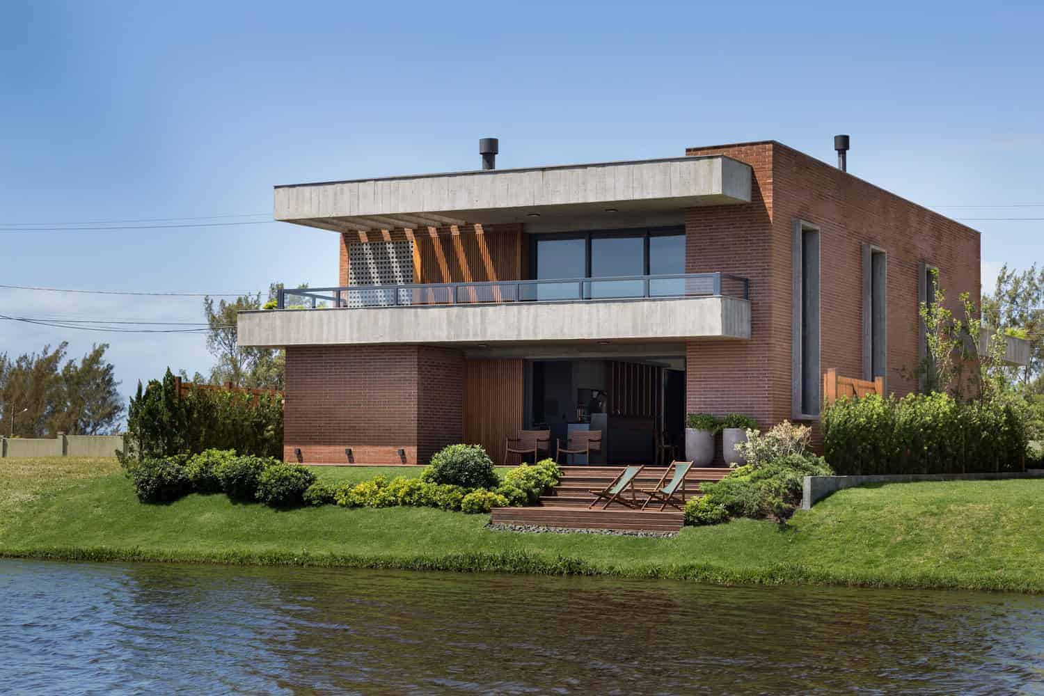 Contemporary modern brick house with spacious balconies, large windows, and lush landscaping near a water body. Stylish architectural design showcasing innovative residential architecture ideas.