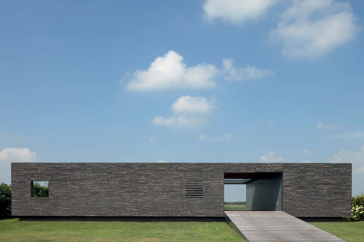Contemporary minimalist black brick modern house with rectangular design and open entrance, surrounded by green lawn, under a clear blue sky with white clouds, showcasing innovative architecture.