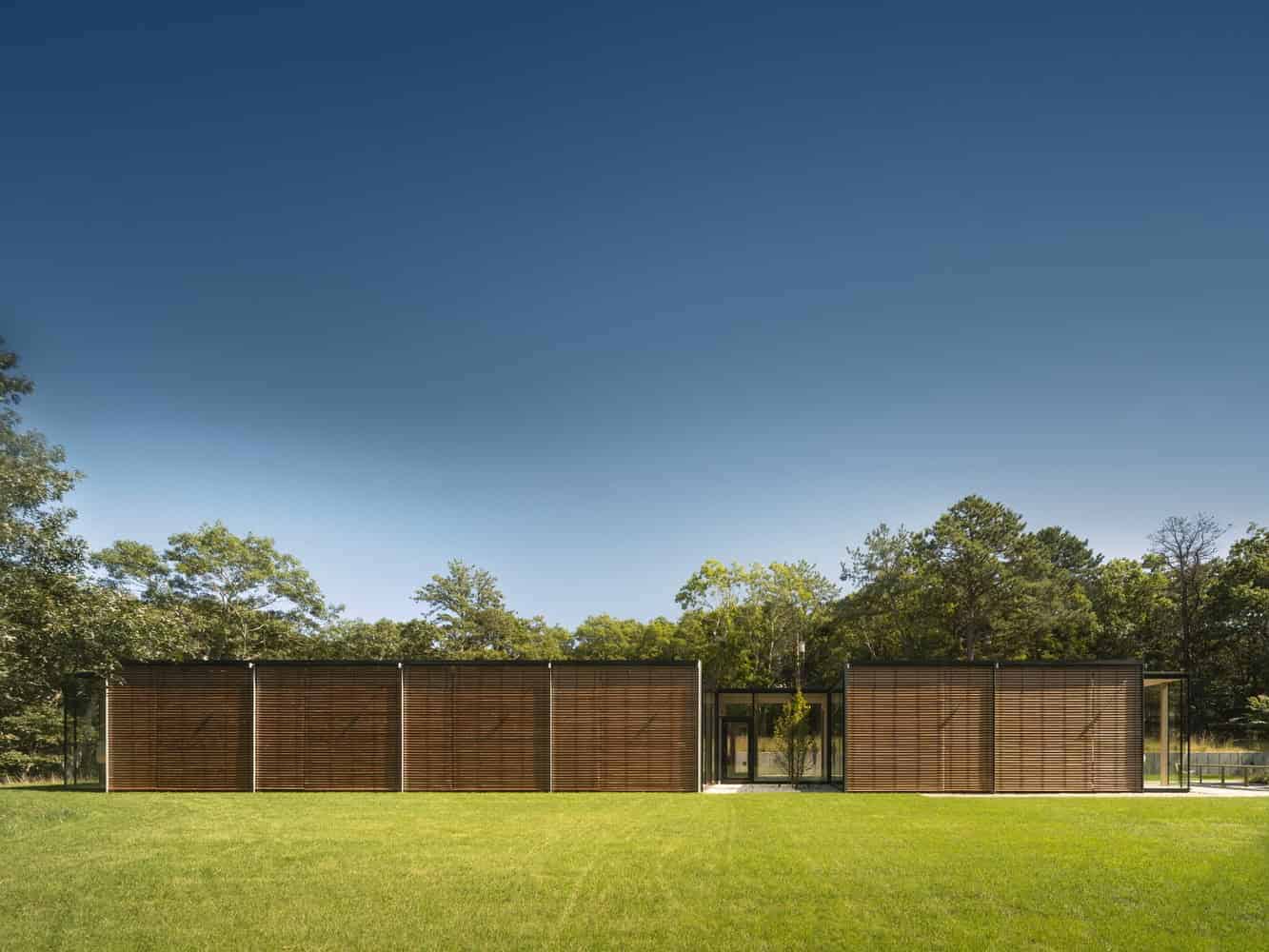 Modern minimalist wooden house with sleek lines and large glass windows, set amidst lush greenery and open lawn, exemplifying contemporary architecture and innovative design.