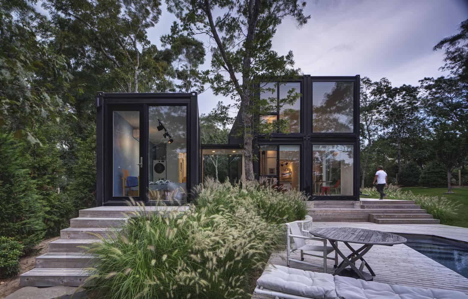 Modern glass house with black steel framing surrounded by lush greenery and mature trees. Features large windows, outdoor seating area, and swimming pool, blending contemporary design with natural landscape.