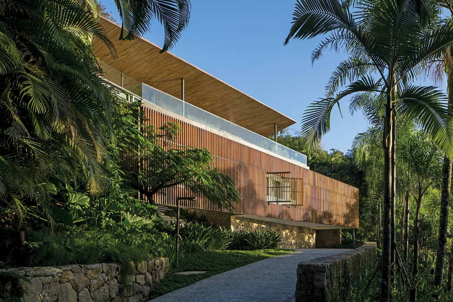 Modern tropical house with wooden slat facade, glass balcony, and lush landscaping, blending contemporary architecture with nature for stylish outdoor living.