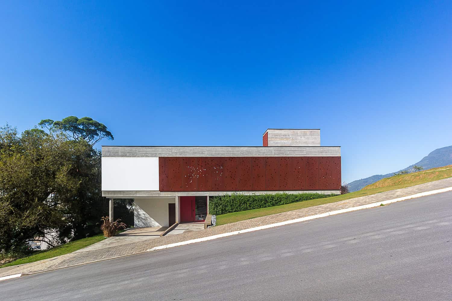 Contemporary modern house with minimalist architectural design, clean lines, and geometric structure on a sloped terrain under a bright blue sky.