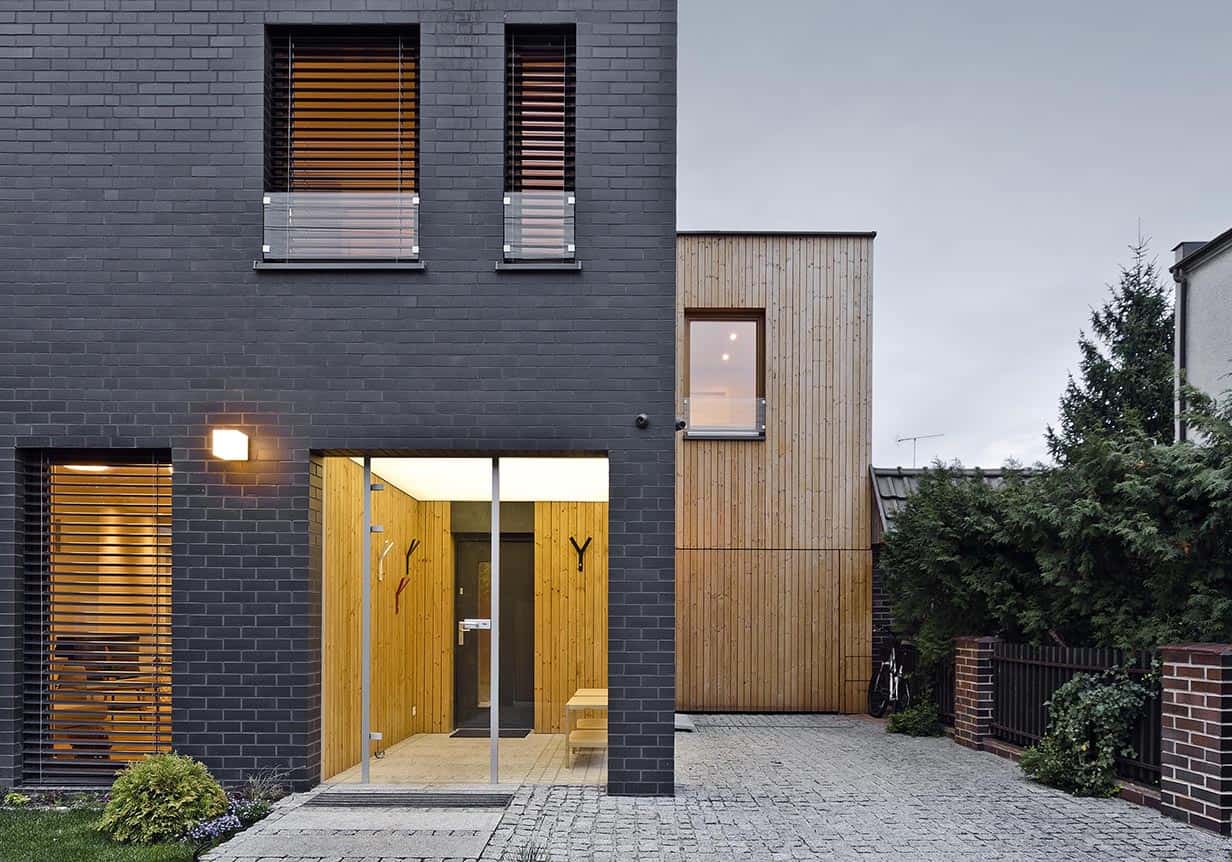 Contemporary house with black brick and natural wood exterior, featuring modern architectural design, large windows, and minimalist entrance in an urban residential setting.