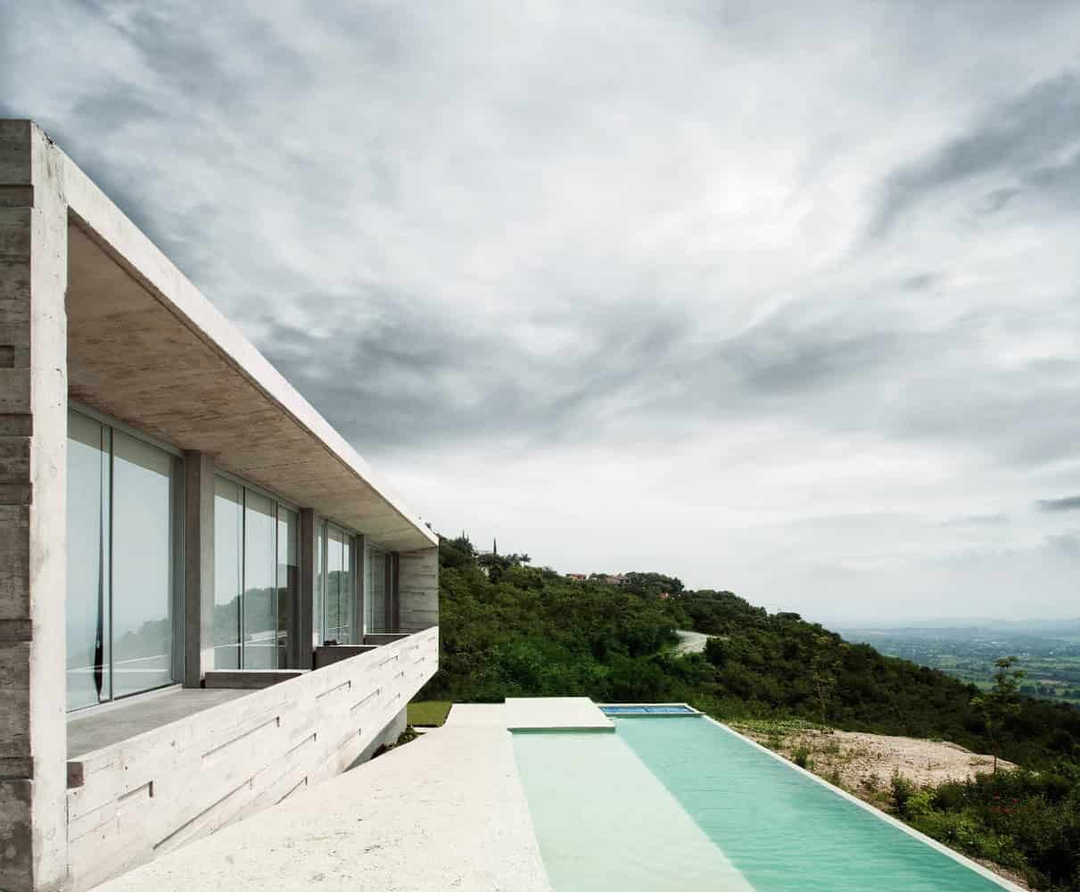 Contemporary modern house with large glass windows, minimalist concrete design, and infinity pool overlooking lush green hillside with cloudy sky.