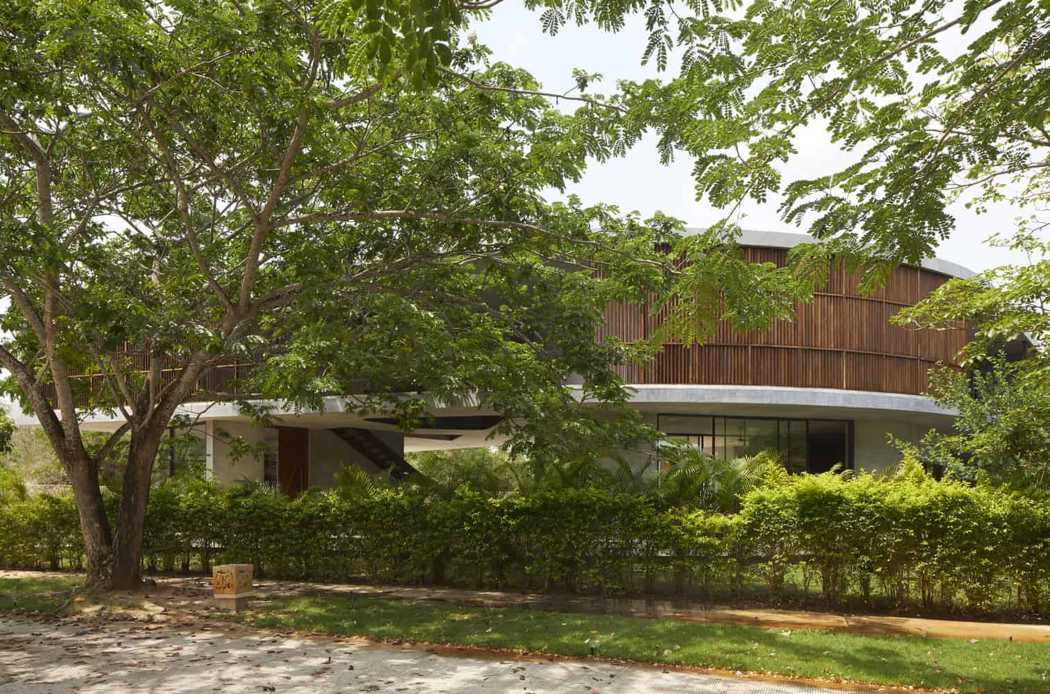 Modern contemporary house with curved wooden facade surrounded by lush green trees and landscaping.