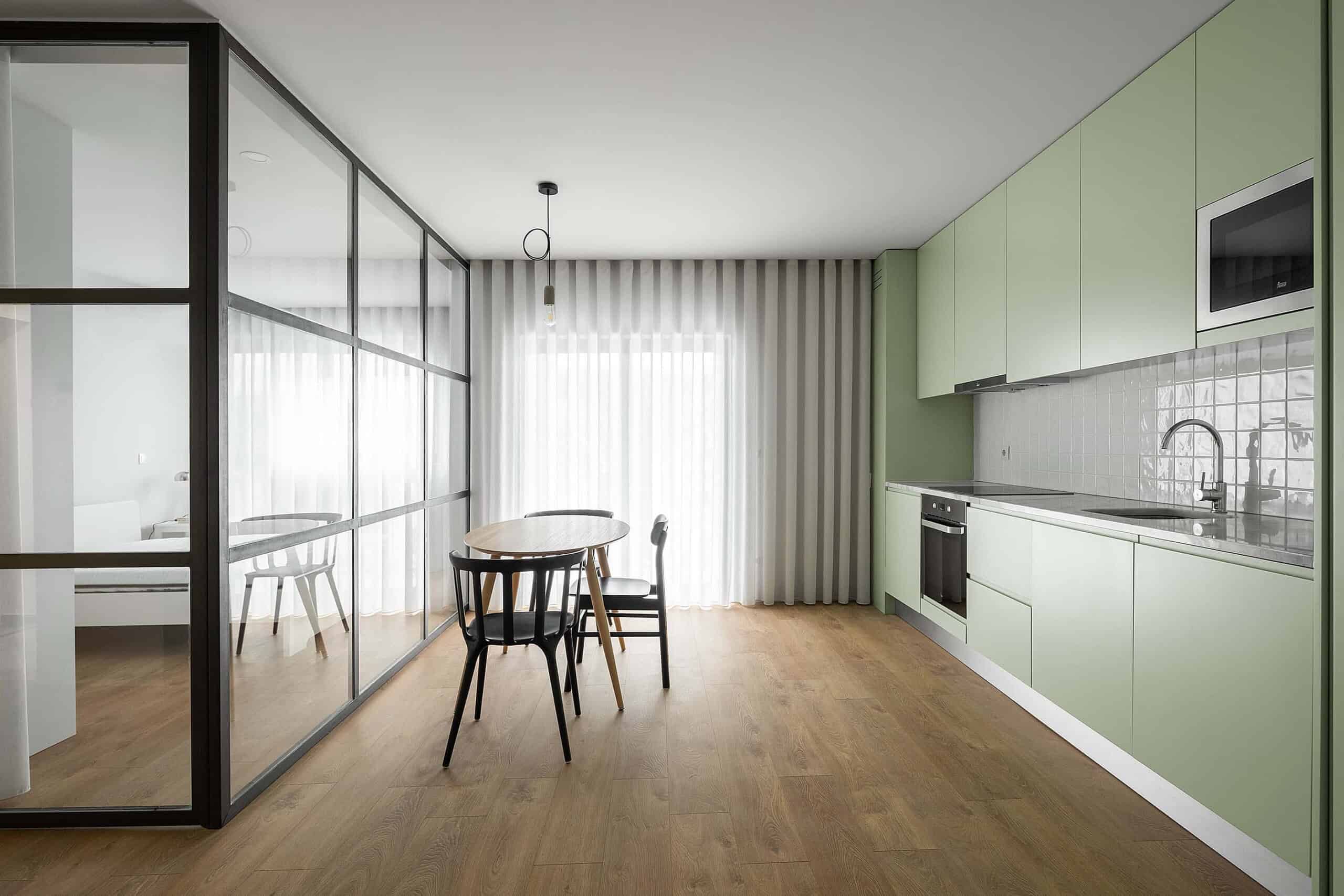 Modern minimalist kitchen with light green cabinetry and open-concept design, featuring wood flooring, sleek appliances, and a glass partition separating dining and living areas.