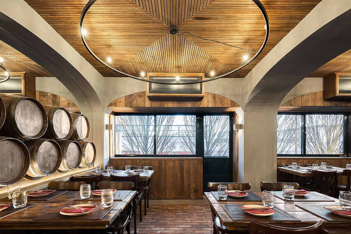 Rustic restaurant interior with wooden wine barrels, brick flooring, arched windows, and warm wood accents, showcasing cozy and stylish architecture design.