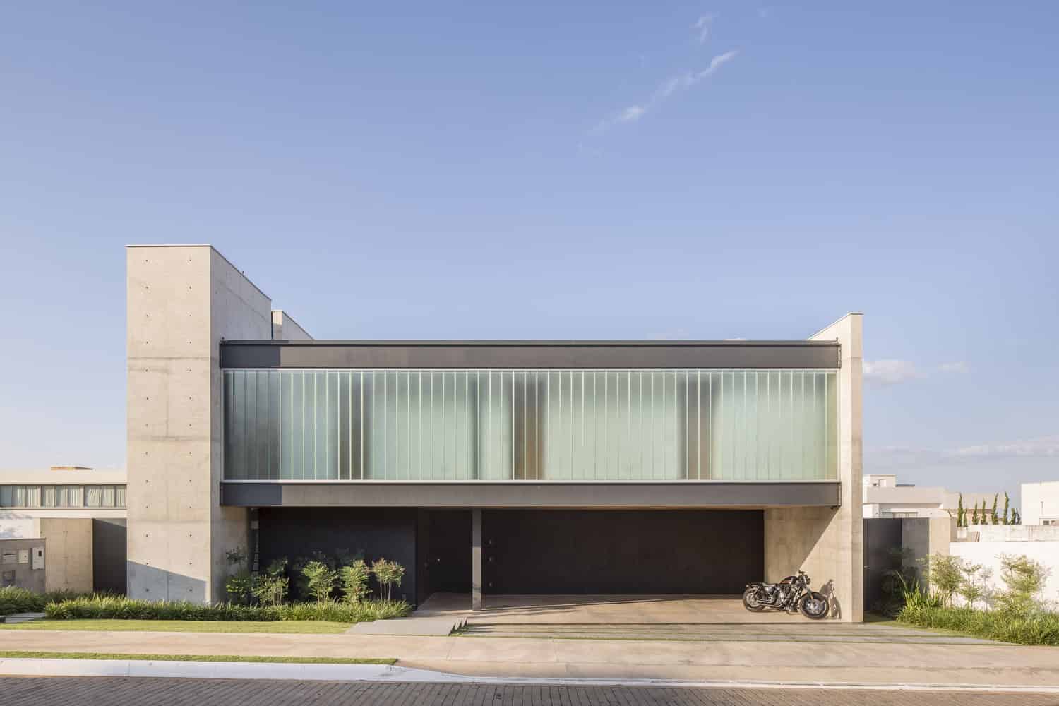 Modern minimalist house with concrete walls and large glass windows, featuring a sleek garage and surrounding greenery.