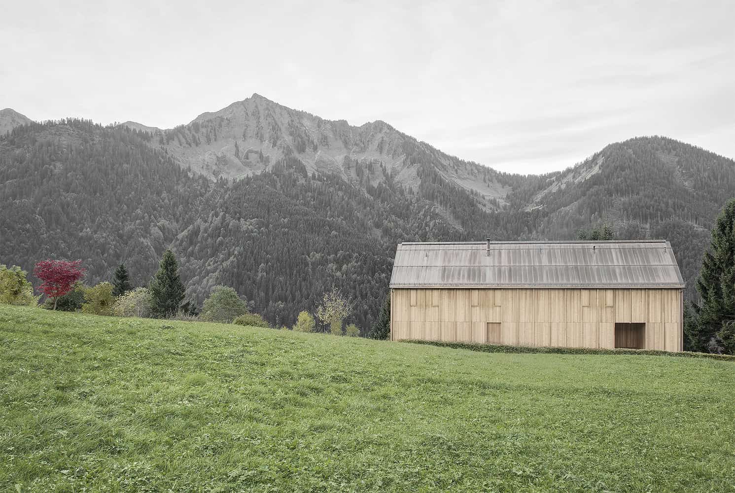 Simple wooden barn situated on green meadow with mountainous landscape in the background, showcasing rustic architecture and natural design elements.