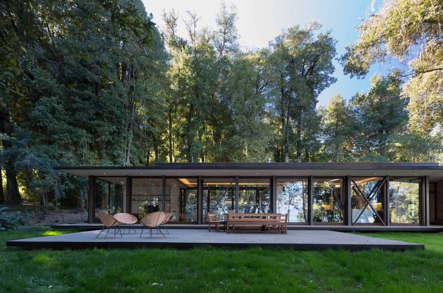 Modern glass house with flat roof nestled in lush forest setting, featuring outdoor patio and contemporary architecture, perfect for nature-inspired luxury living.
