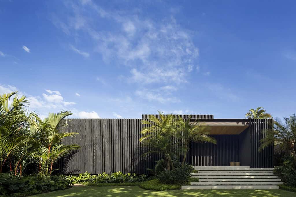 Modern minimalist black house with wooden slat exterior, surrounded by lush tropical landscaping and palm trees, under a clear blue sky.