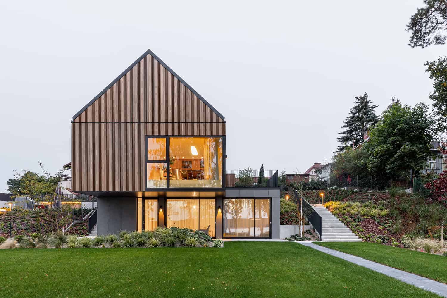 Contemporary modern house with sleek exterior design featuring vertical wooden panels and expansive glass windows, set in a lush landscape with manicured lawn and outdoor lighting, showcasing innovative architecture.