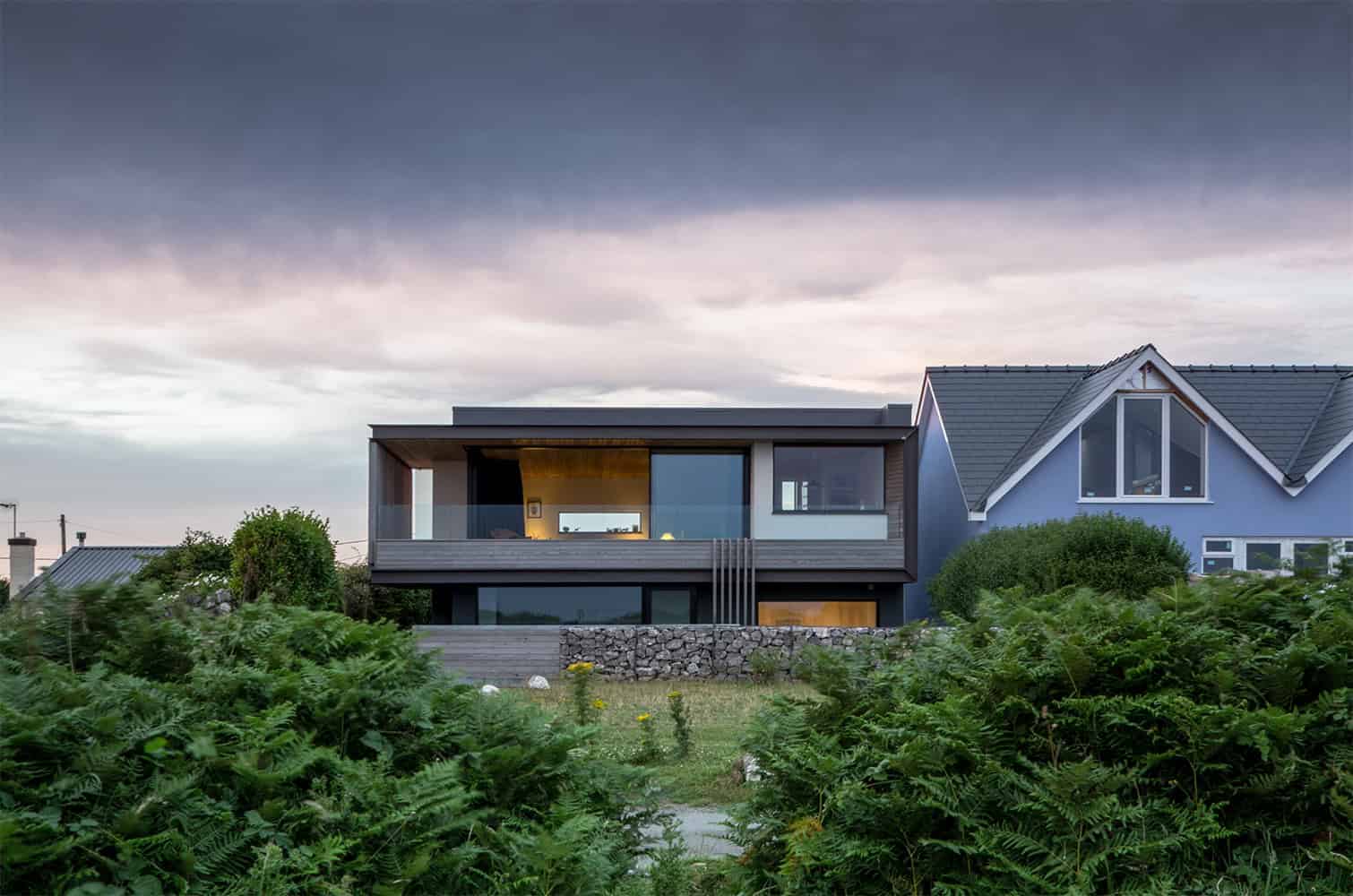 Modern contemporary house with sleek design, large glass windows, and unique architectural features, set against a lush green landscape and a dramatic evening sky.