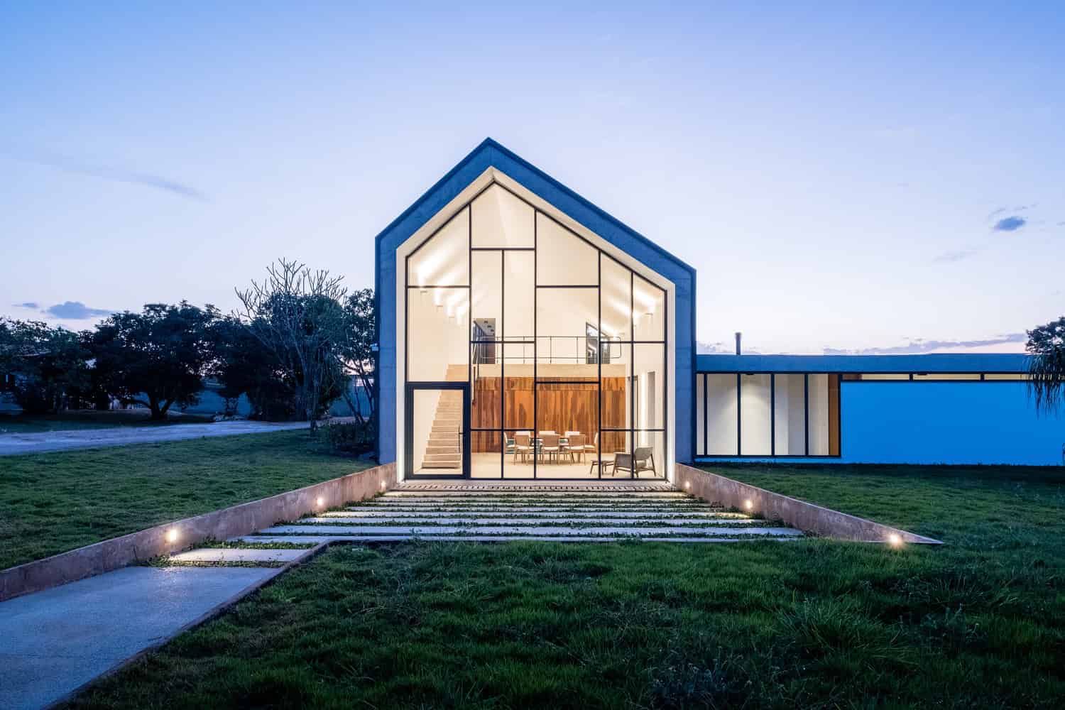 Contemporary modern house with large glass windows, minimalist design, and open interior view, set in landscaped garden during twilight.