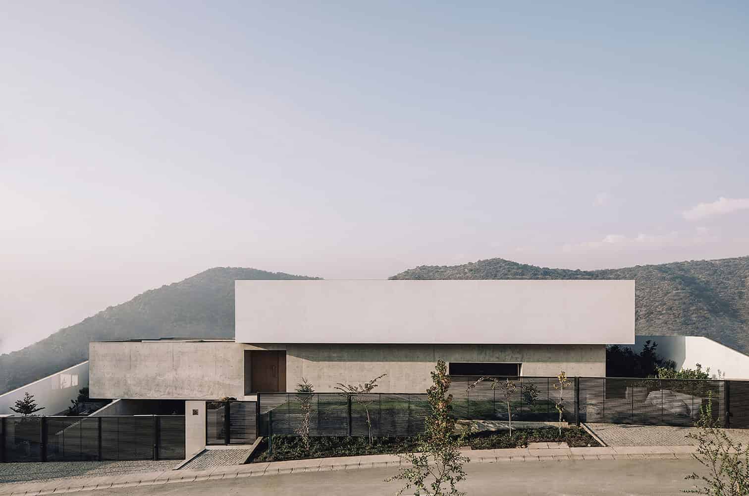 Modern minimalist concrete house with clean lines and geometric design, set against scenic mountain backdrop, surrounded by a sleek metal fence and landscaped garden.