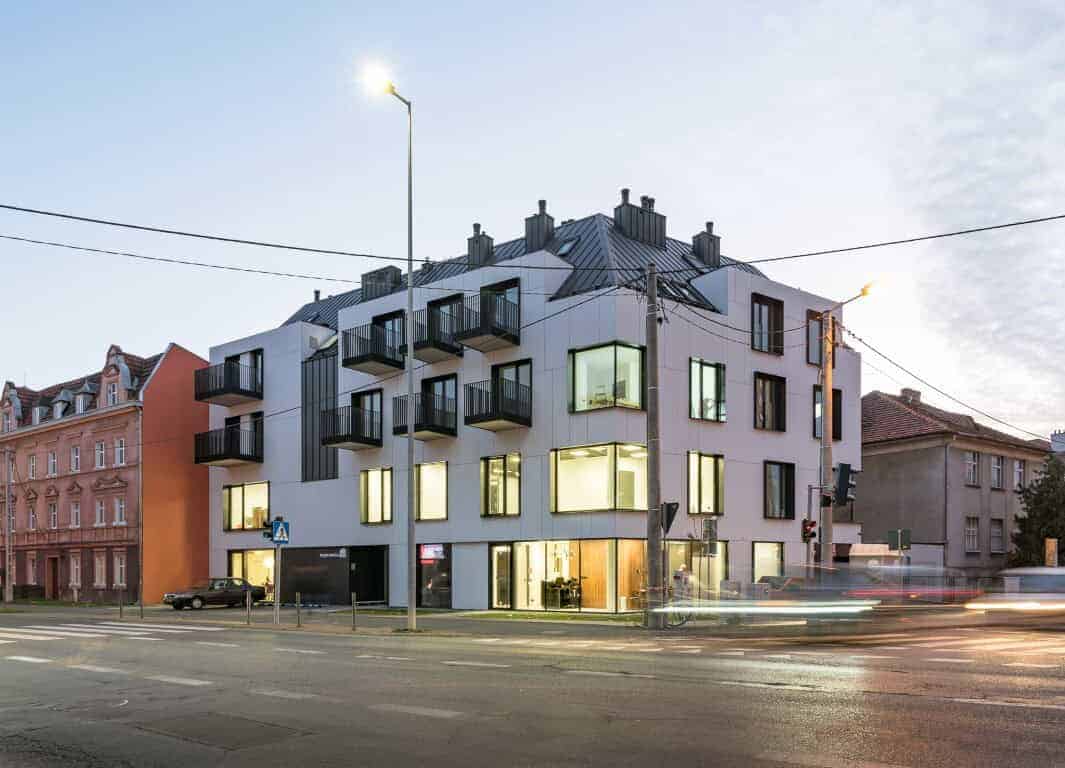 Contemporary modern residential building with geometric design, large glass windows, and small balconies, located on city street at dusk, emphasizing innovative architectural art and urban living spaces.
