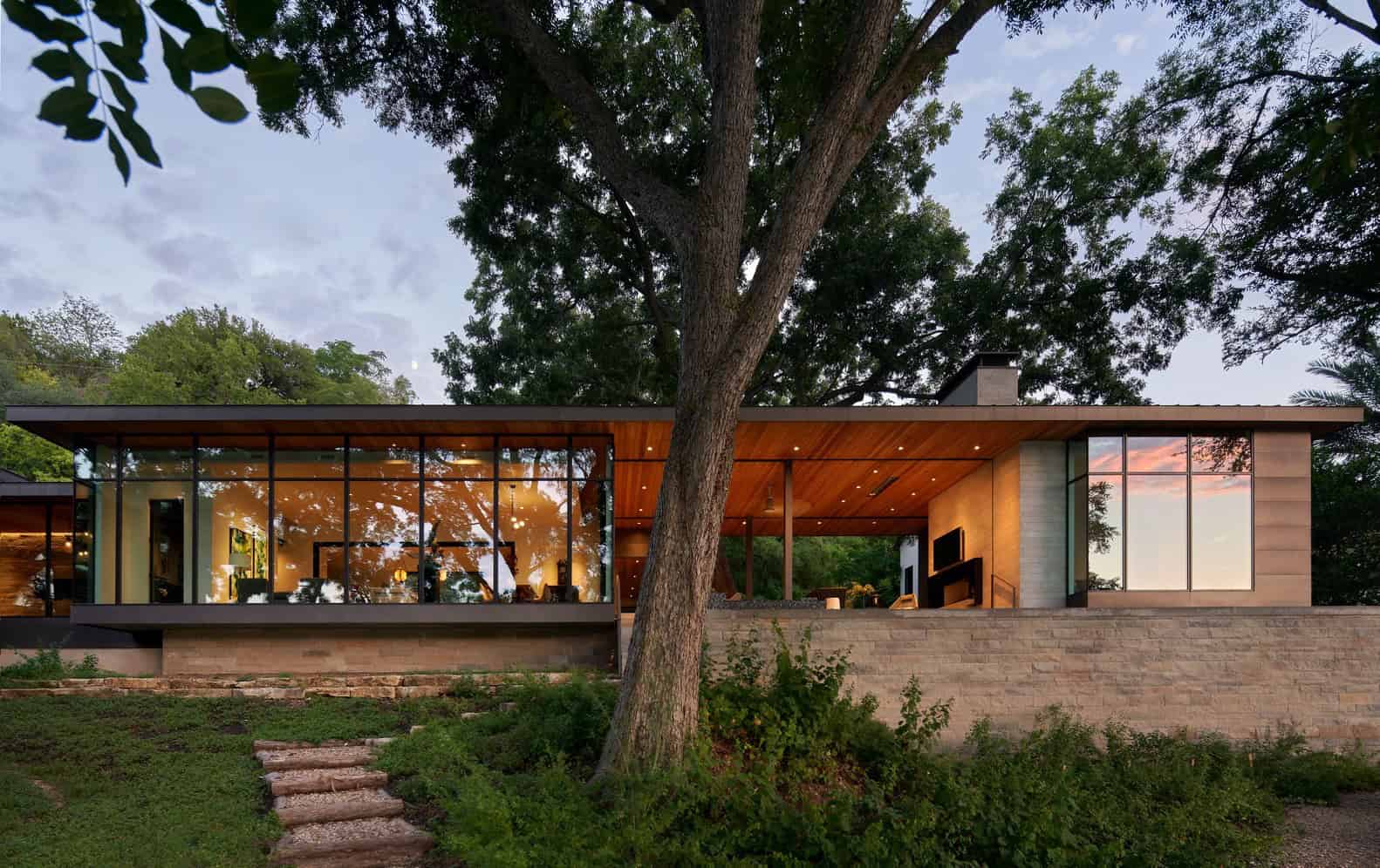 Sustainable modern house with extensive glass walls, natural wood accents, and integrated indoor and outdoor living spaces amidst lush greenery and mature trees.