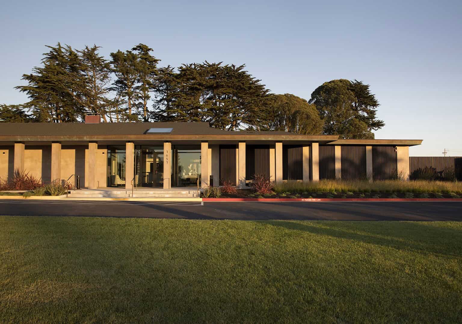 Modern single-story commercial building with large glass entrance, concrete columns, and sleek black vertical louvers, set amidst lush landscaping and tall trees under clear skies.