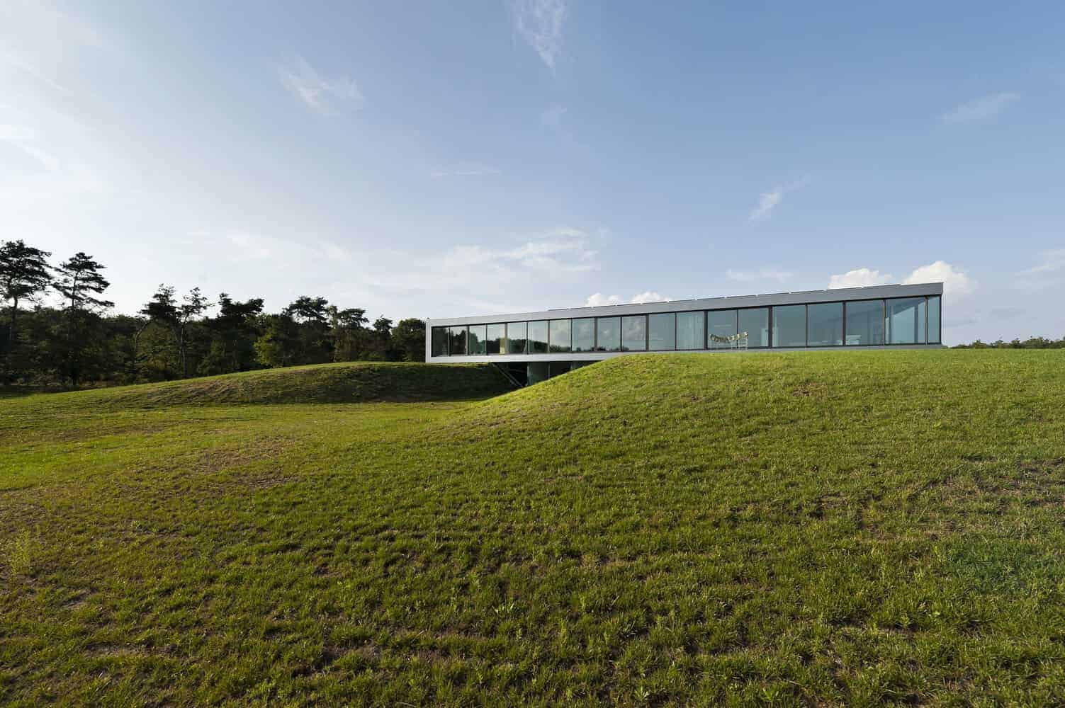 Sleek modern glass house situated on a grassy hillside with lush trees in the background, showcasing contemporary architecture and landscape integration.