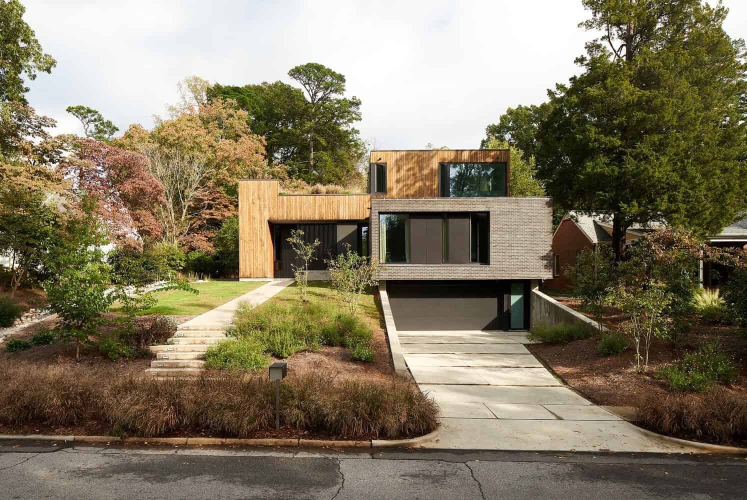 Modern contemporary house with unique geometric design, combination of wood, brick, and large glass windows, set in lush landscaped surroundings.