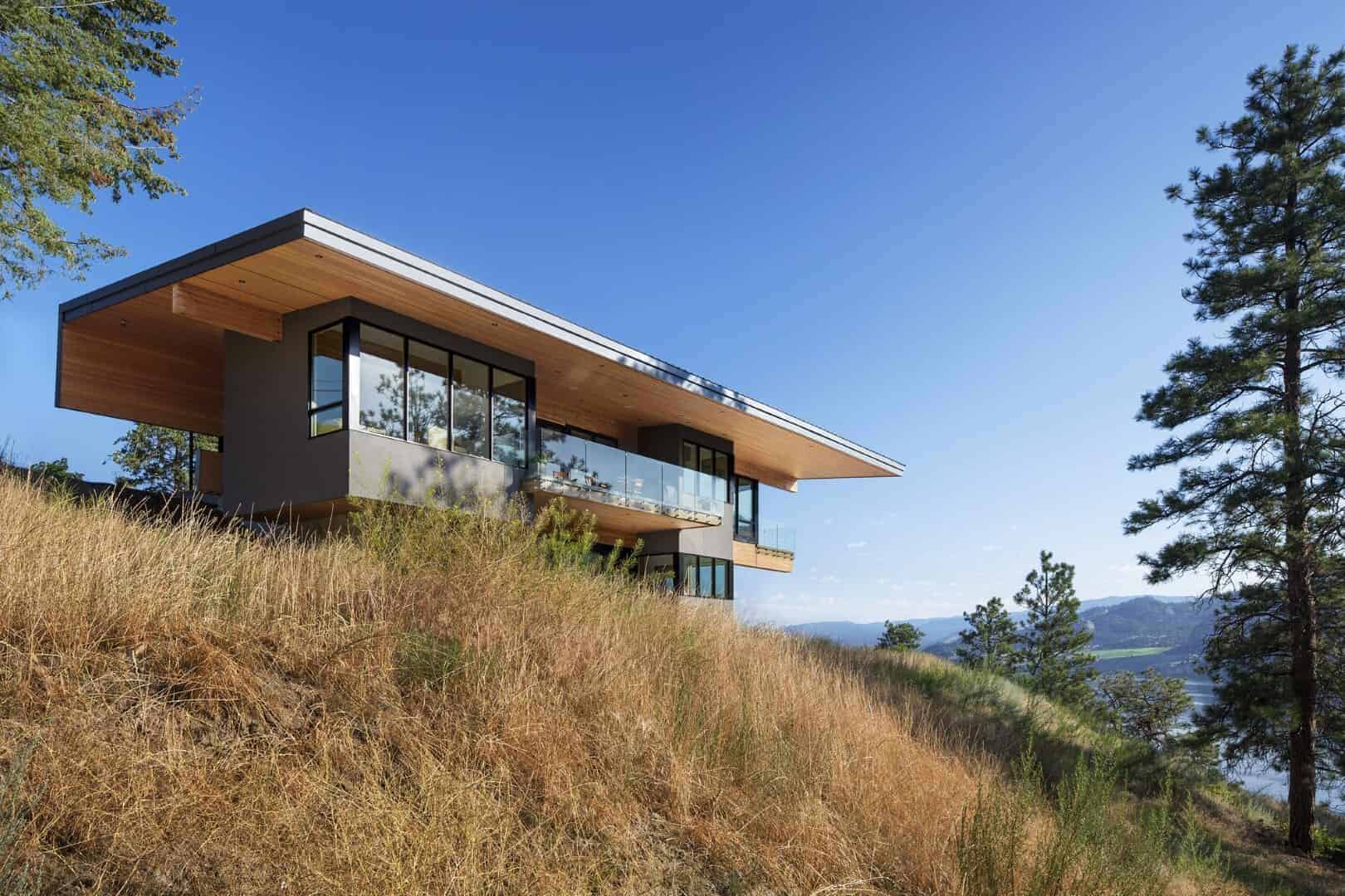 Modern hillside house with extensive glass windows, wooden accents, and a flat roof, set against a scenic mountain landscape for contemporary architecture and luxury home design.