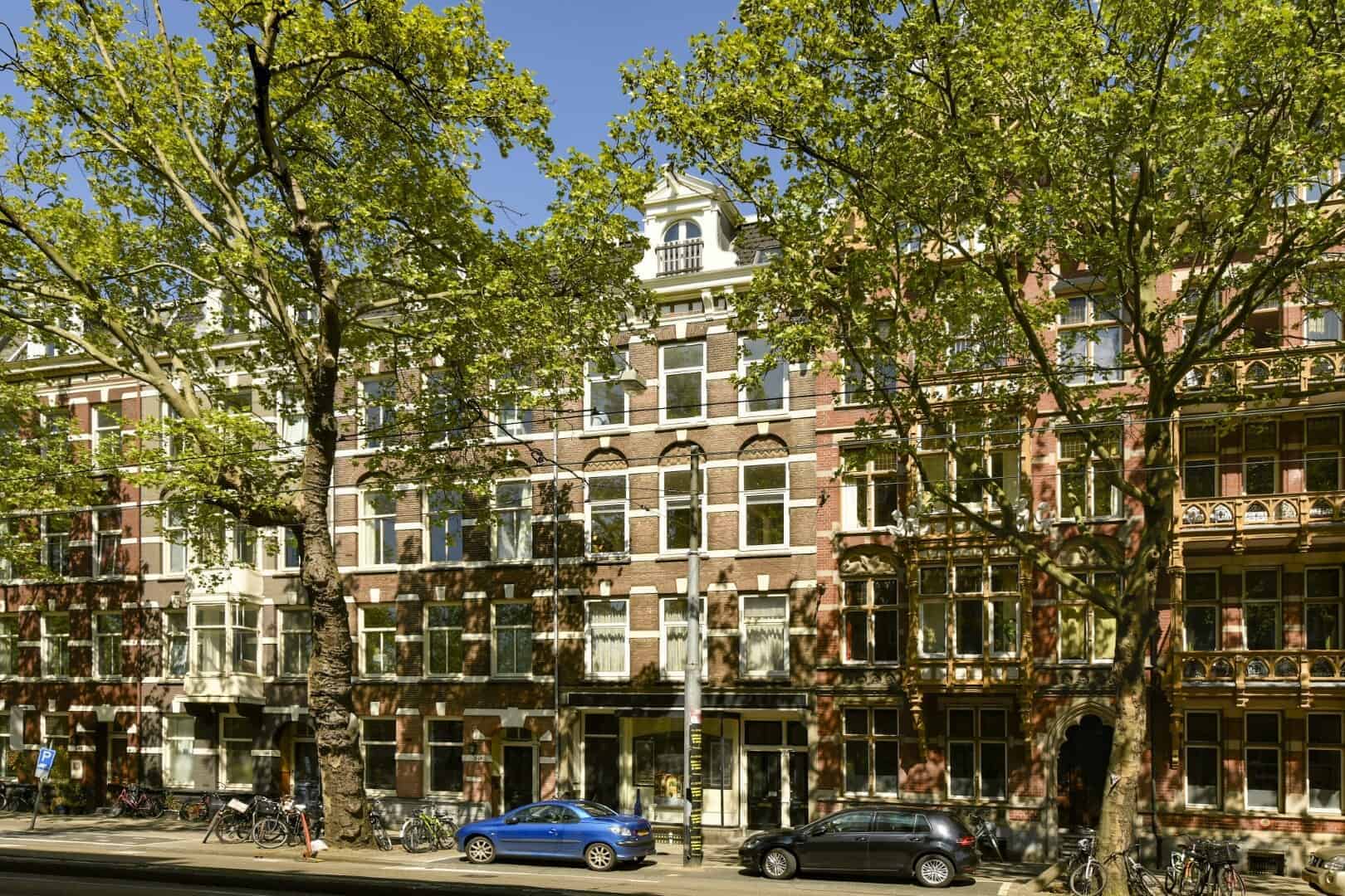 Elegant historic Amsterdam canal house with traditional Dutch architecture, large windows, ornate gabled roof, surrounded by lush green trees in a lively cityscape setting.