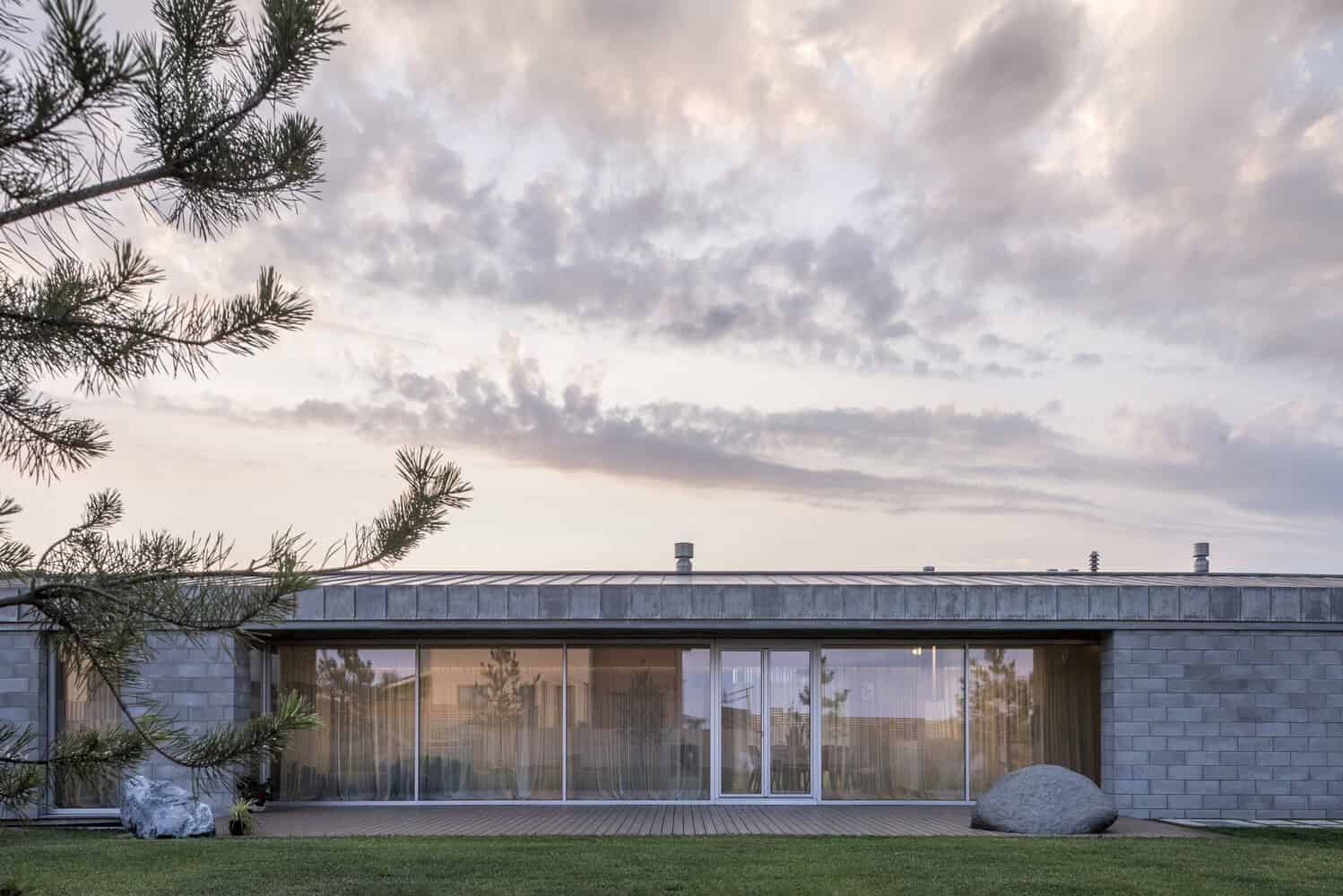 Contemporary modern house with large glass windows, metal roof, and stone exterior surrounded by a well-maintained lawn and natural landscape.