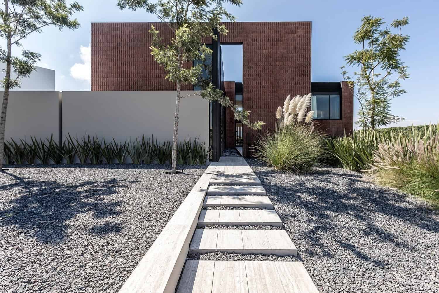 Contemporary minimalist house with modern architectural design and clean lines, featuring red brick exterior, large windows, and landscaped front yard with drought-tolerant plants and a stone walkway.