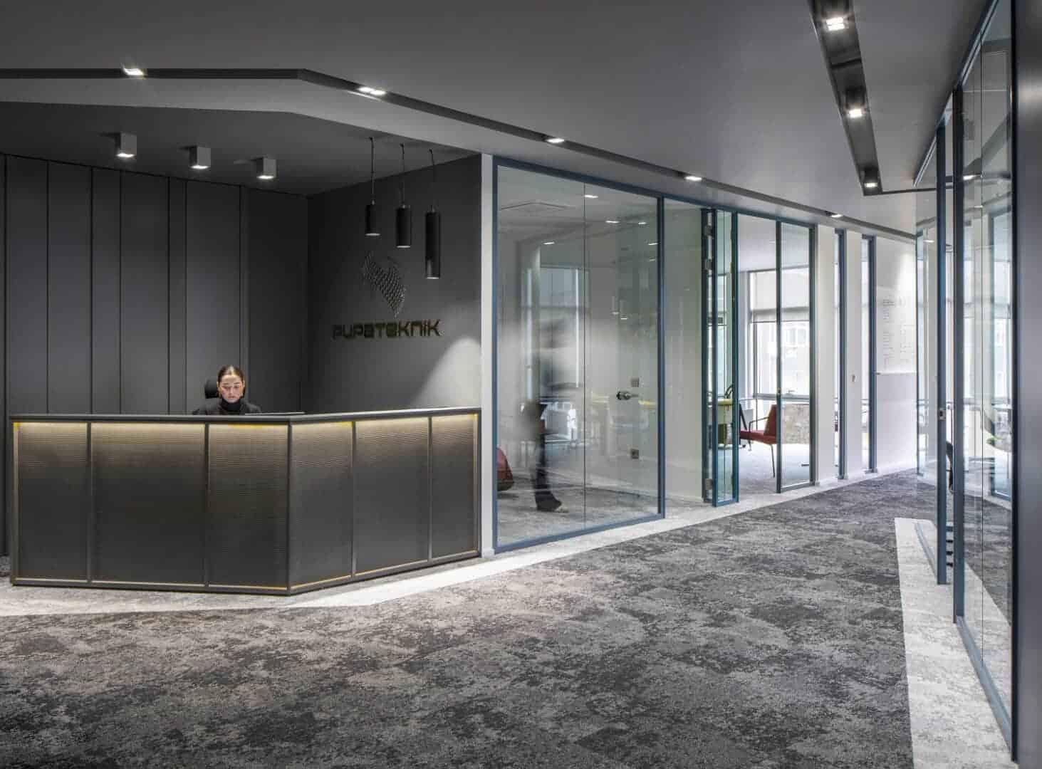 Modern office reception area with sleek dark gray wall panels, stylish lighting fixtures, and glass partition walls creating an open and contemporary workspace environment.