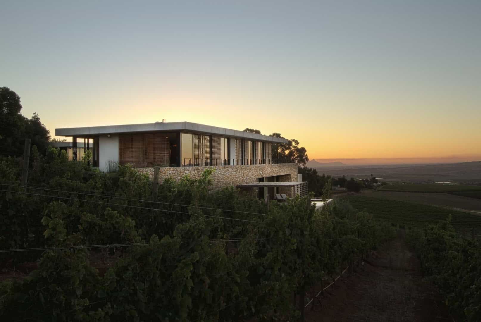Contemporary hillside house with modern architecture, large glass windows, and a stone exterior, overlooking vineyards at sunset, showcasing innovative design and integration with natural surroundings.