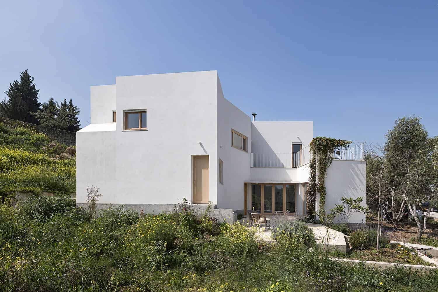 Modern minimalist white house with geometric architectural design and large windows, surrounded by lush greenery and garden landscape.
