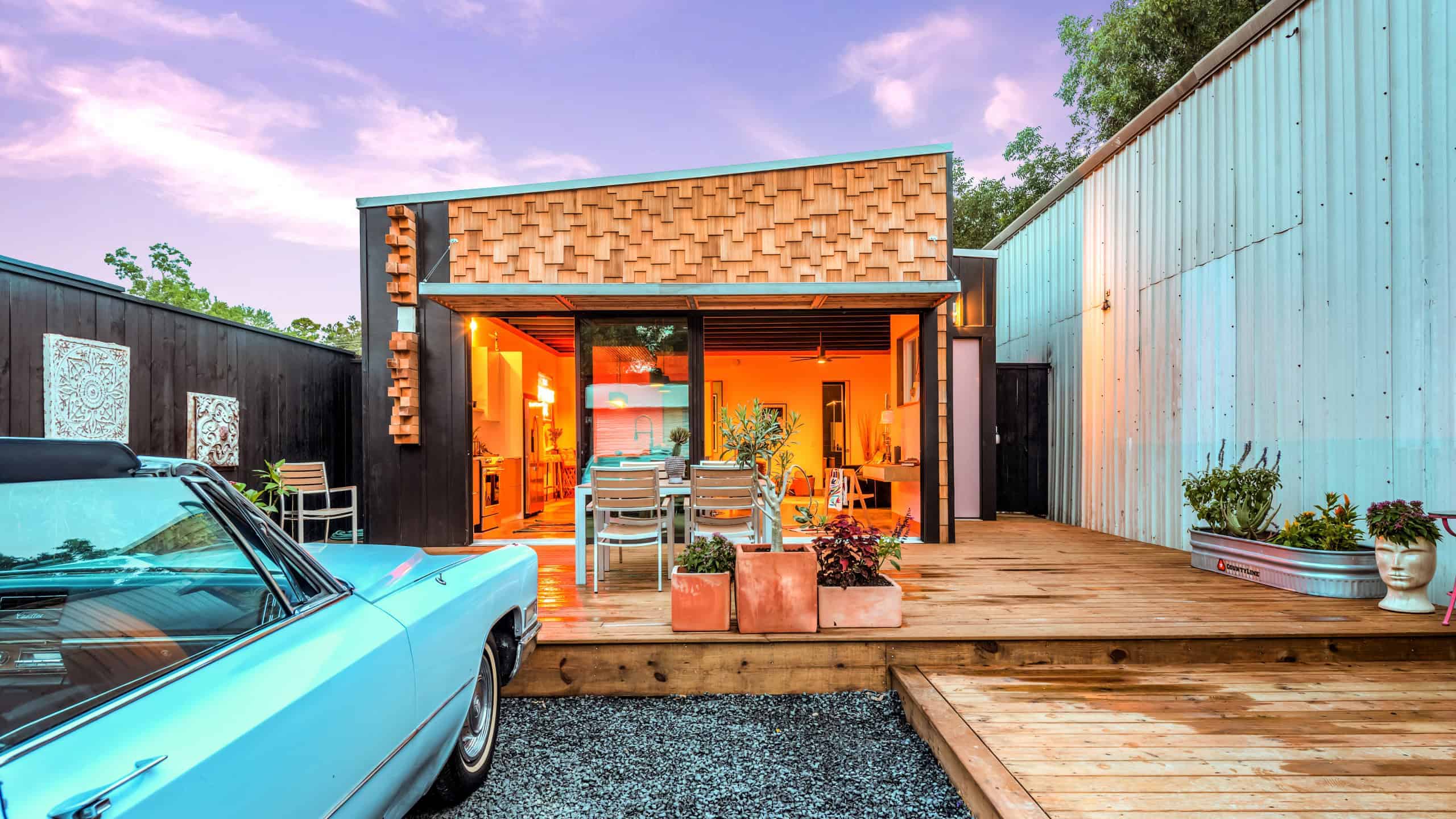 Modern tiny house with a wooden deck, contemporary exterior design, and vibrant interior lighting, surrounded by greenery and vintage car parked outside.