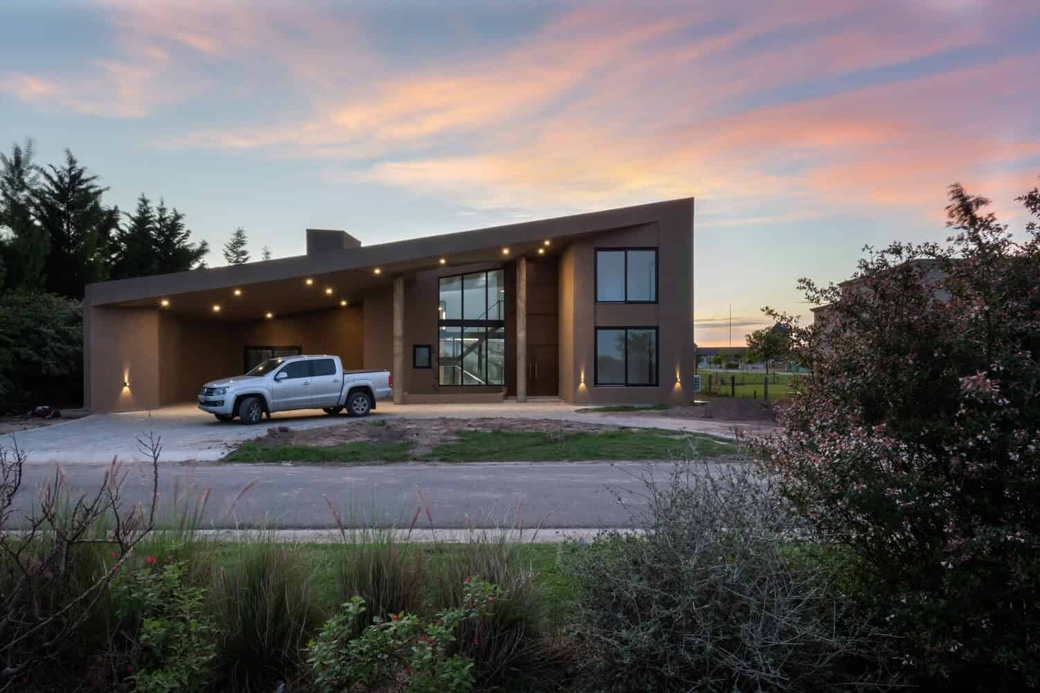 Modern contemporary house with sleek architectural design, large glass windows, and spacious driveway, illuminated by warm exterior lighting at sunset.