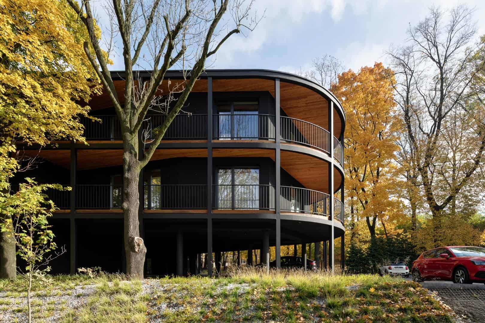 Contemporary black and wood multi-story residential building with curved balconies surrounded by autumn trees and parked cars.