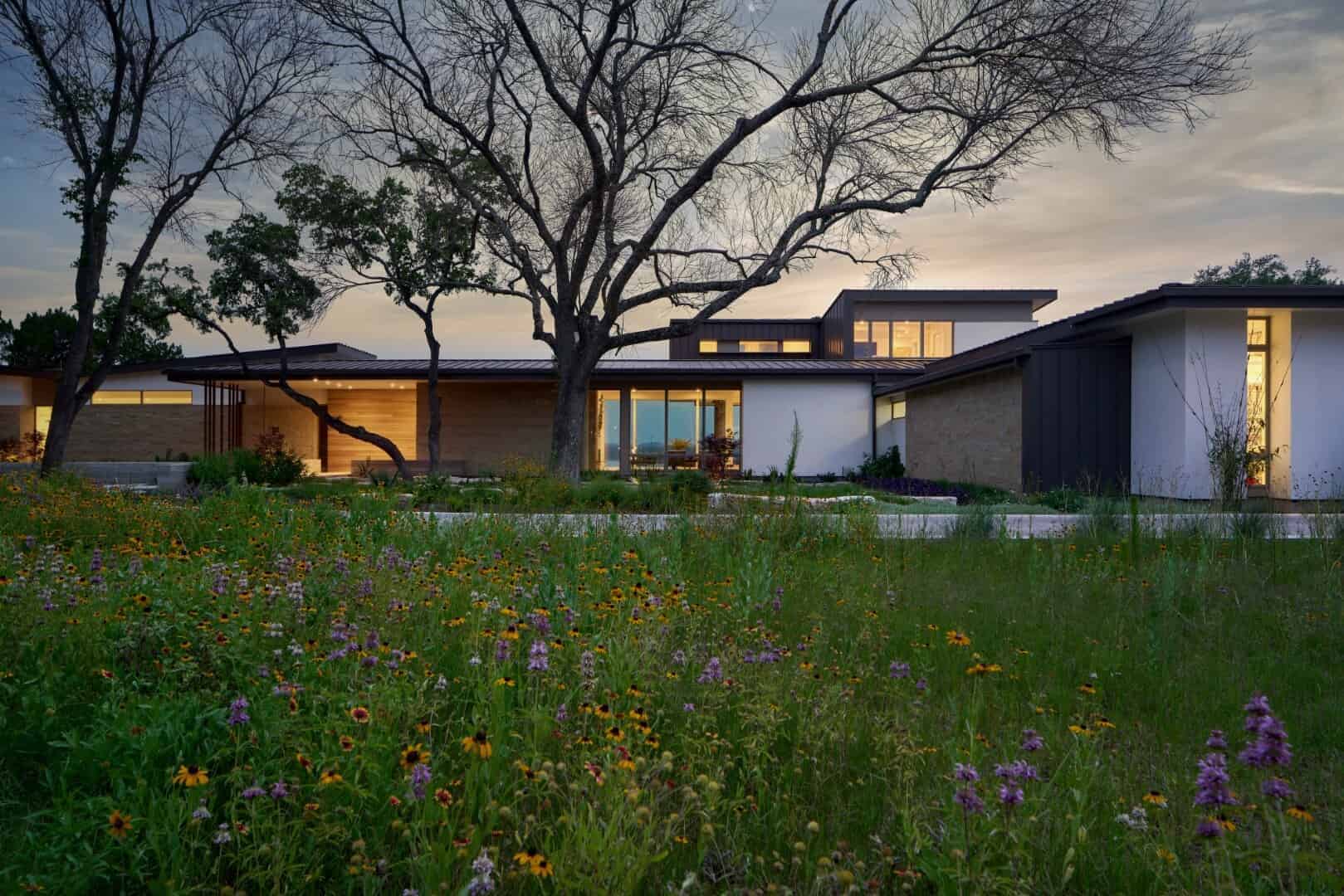 Modern contemporary house with sleek design and large windows, surrounded by a colorful flower garden and mature trees, showcasing innovative architecture and landscape design.