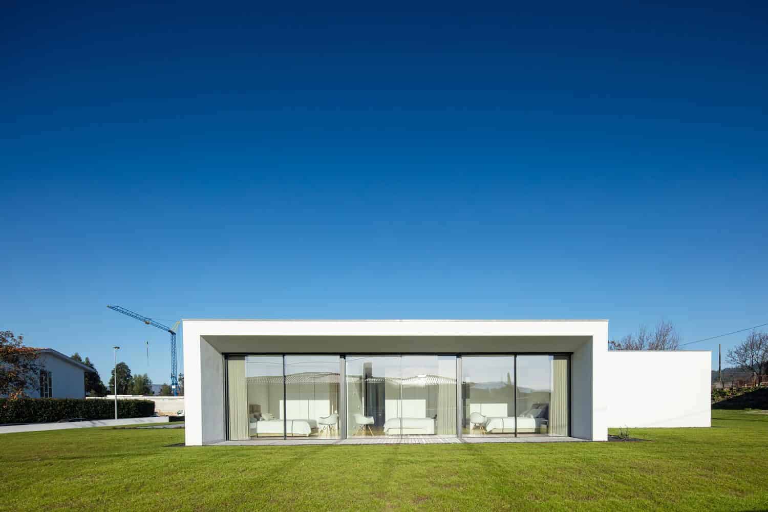 Contemporary minimalist modern house with large glass sliding doors and flat roof, surrounded by lush green lawn under clear blue sky.
