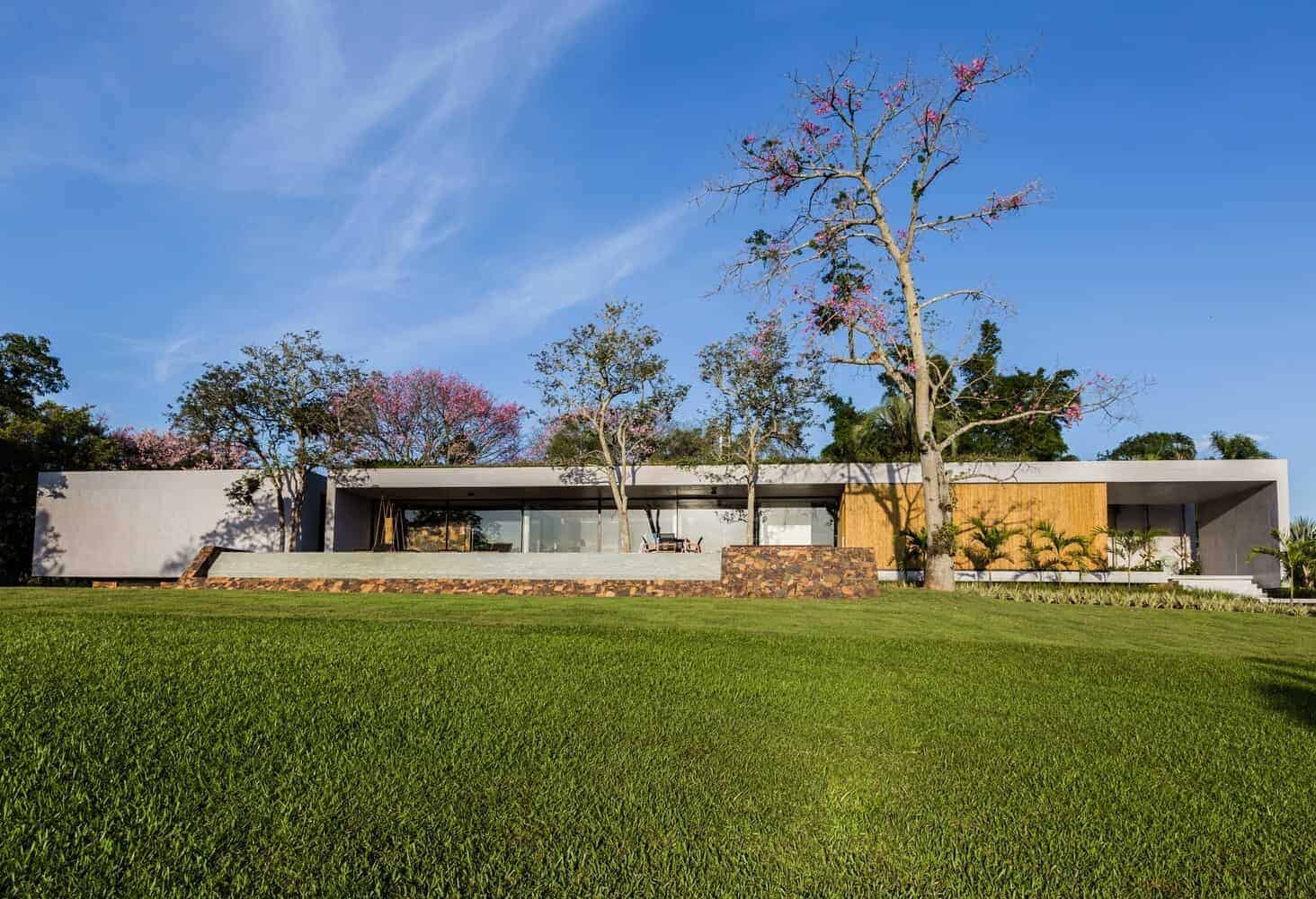Modern minimalist house with clean lines and large glass windows in a lush green landscape under a clear blue sky.