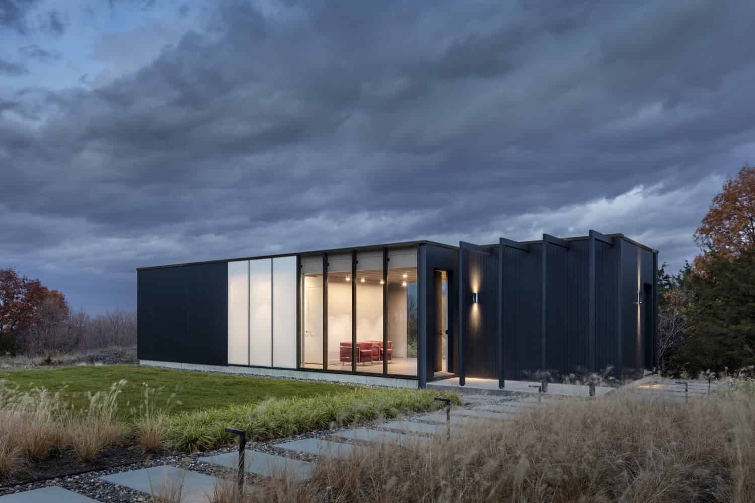 Sleek modern black minimalist house with large glass windows and contemporary lighting, set against a dark, cloudy sky and surrounded by natural landscape.