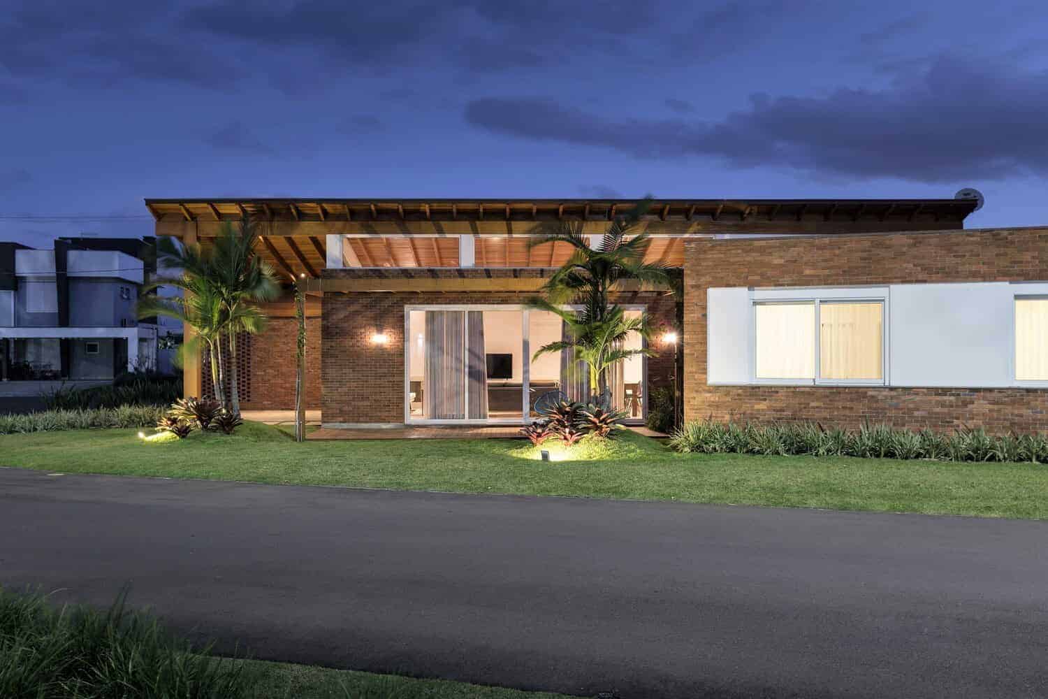 Contemporary modern house with brick and white exterior, illuminated landscape lighting, lush lawn, tropical plants, and large glass sliding doors showcasing sleek architecture at dusk.