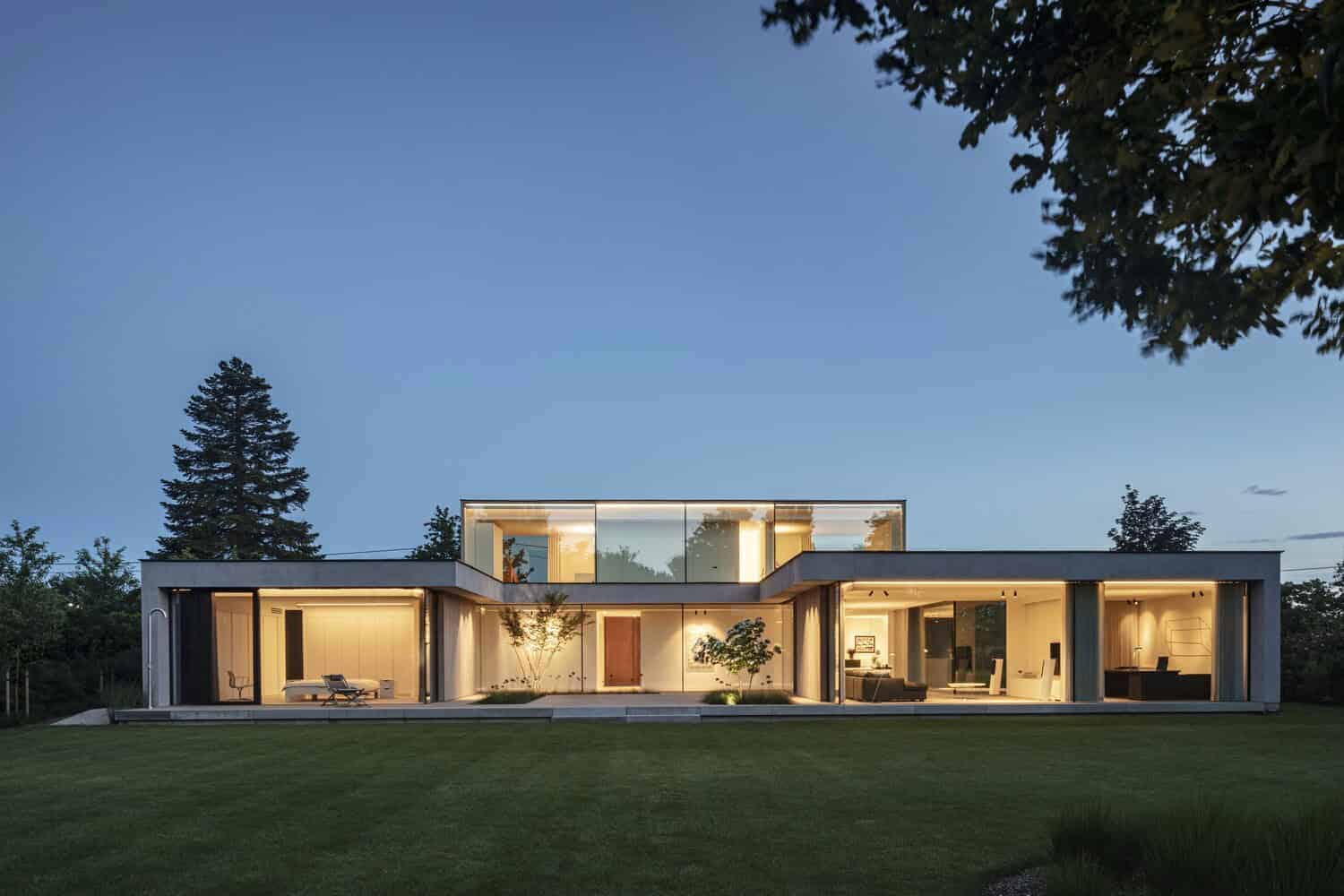 Contemporary minimalist house with sleek glass walls and modern interior design, surrounded by lush greenery and set against a serene evening sky.