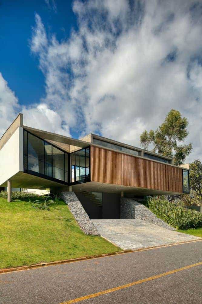 Sleek modern house with cantilevered design, large glass windows, and wooden exterior, set on a grassy hillside under a dramatic sky.
