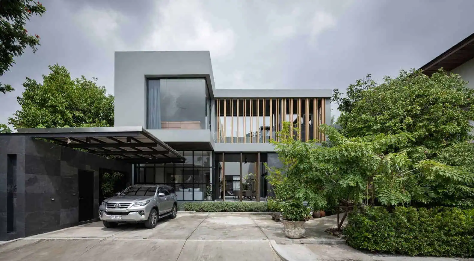 Contemporary modern house with sleek architectural design featuring large glass windows, wooden accents, and a spacious driveway surrounded by lush greenery.