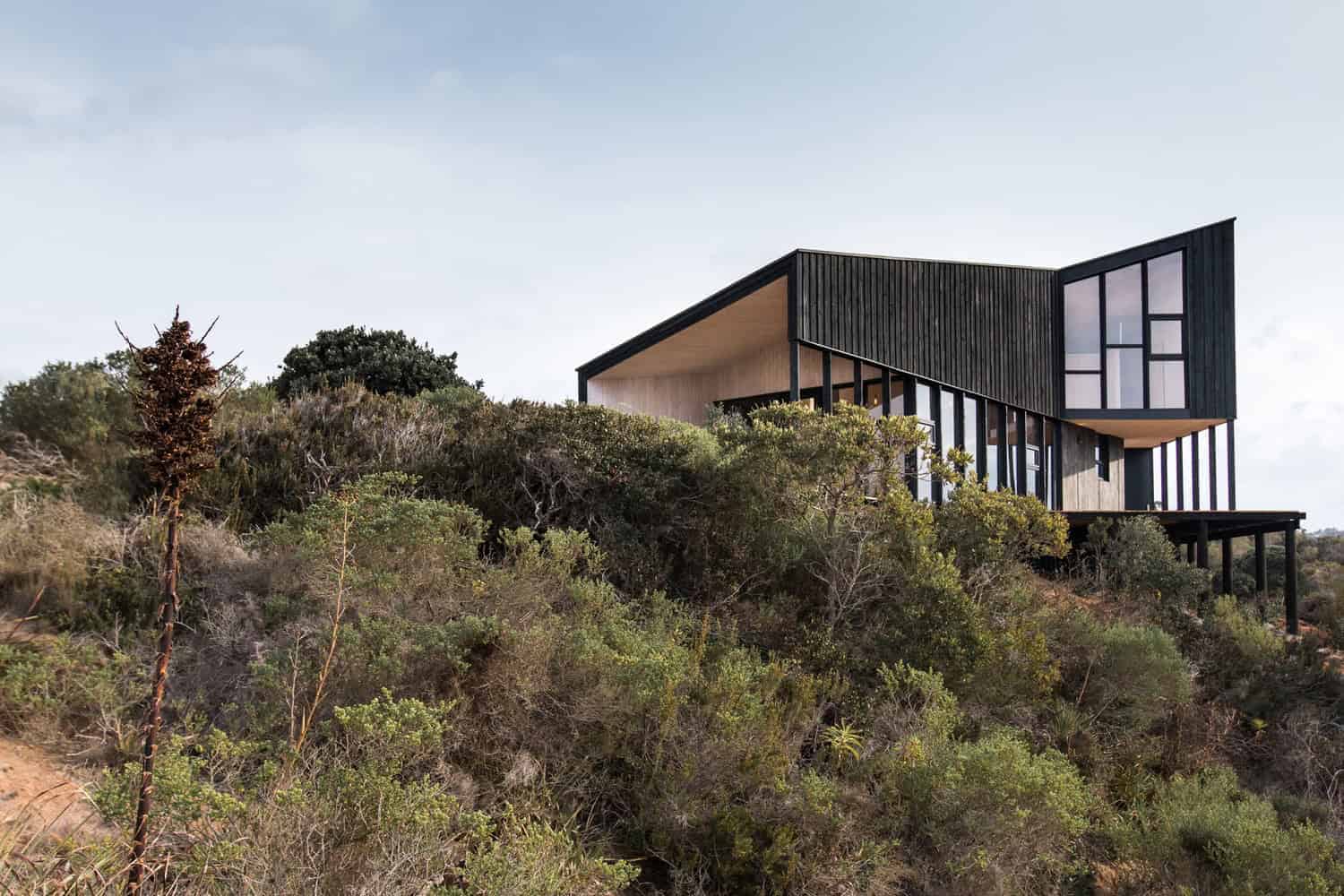 Modern hillside house with black and wood exterior, large windows, and contemporary architectural design amidst lush greenery.