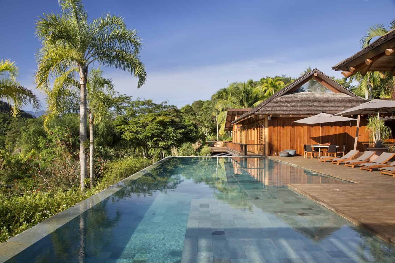 Luxury tropical villa with infinity pool, surrounded by lush greenery and palm trees, perfect for relaxing by the water in a beautiful natural setting.