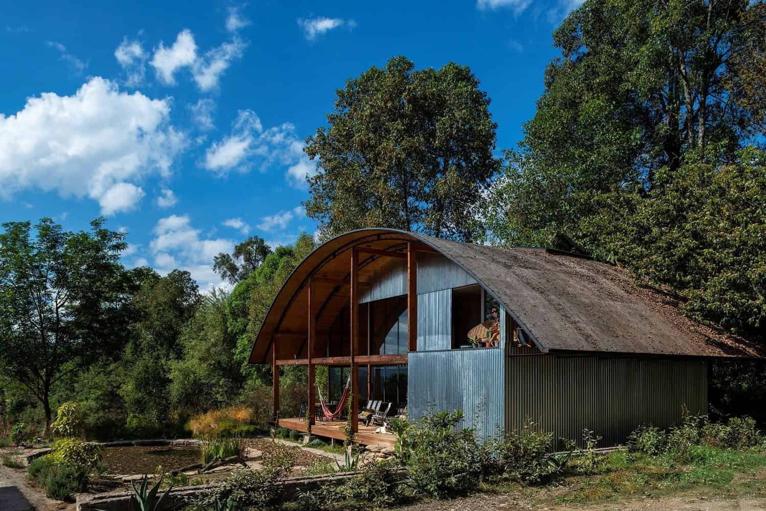 Modern eco-friendly house with curved metal roof surrounded by lush greenery and tall trees, showcasing innovative sustainable architecture and innovative design in nature.