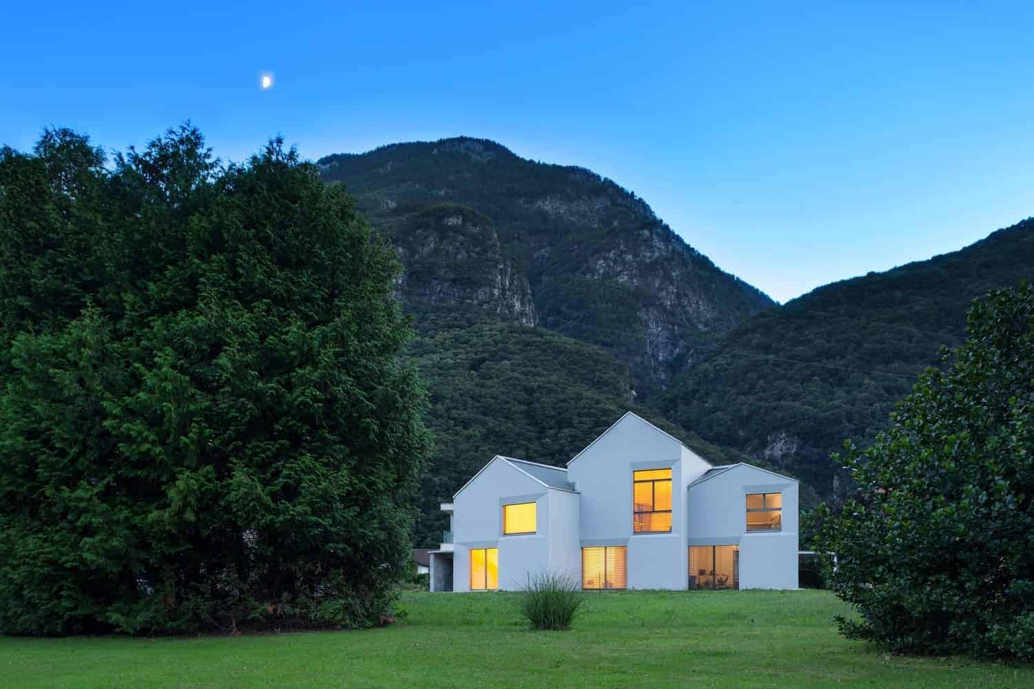 Contemporary modern house with geometric design, large windows, and white exterior, set against lush green landscape and mountain backdrop in scenic outdoor environment.