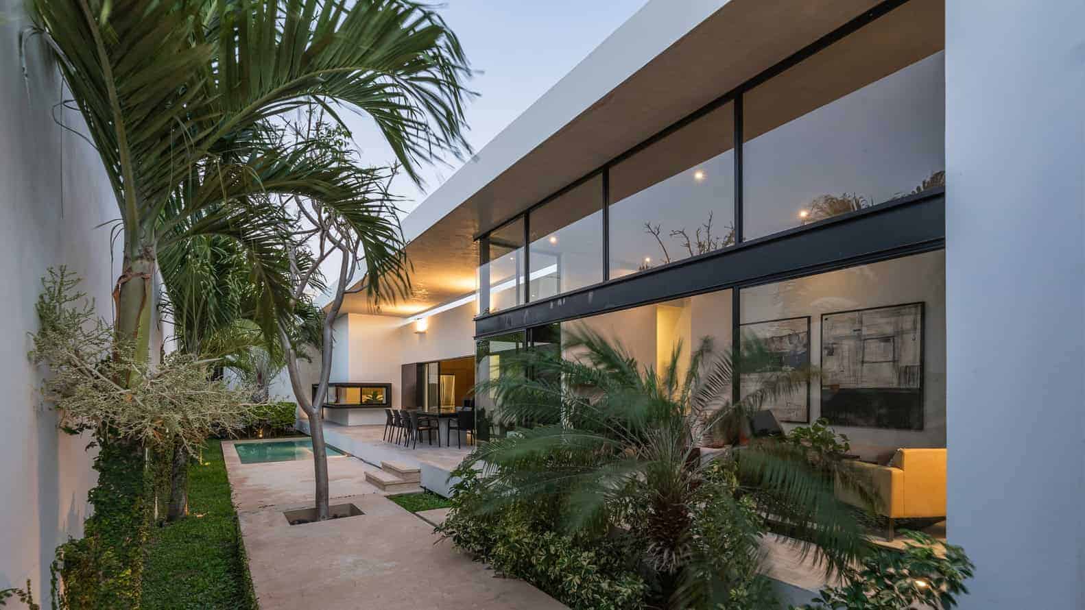 Contemporary modern house exterior with sleek glass walls, lush tropical landscaping, and cozy outdoor seating, highlighting innovative architecture and stylish design elements.