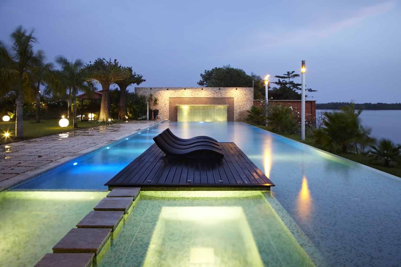 Luxurious modern infinity pool with scenic lake view, wooden deck, ambient lighting, and lush landscaping, showcasing sleek contemporary architecture and outdoor design.